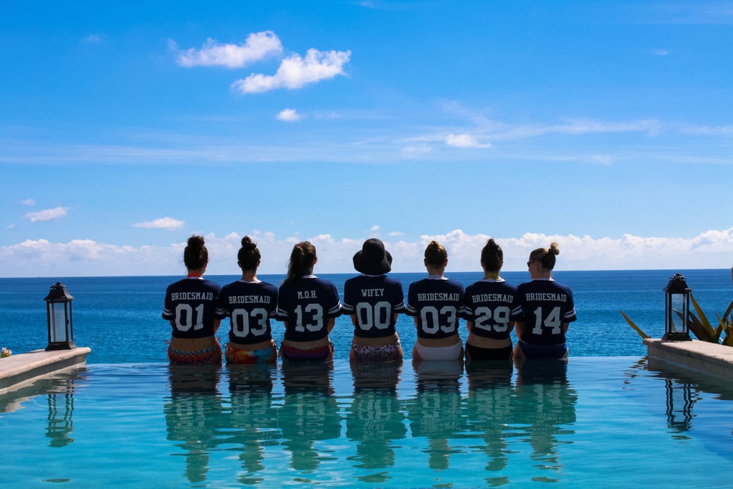 A group at a bachelorette party wearing personalized custom t-shirts line up along the side of a pool. 