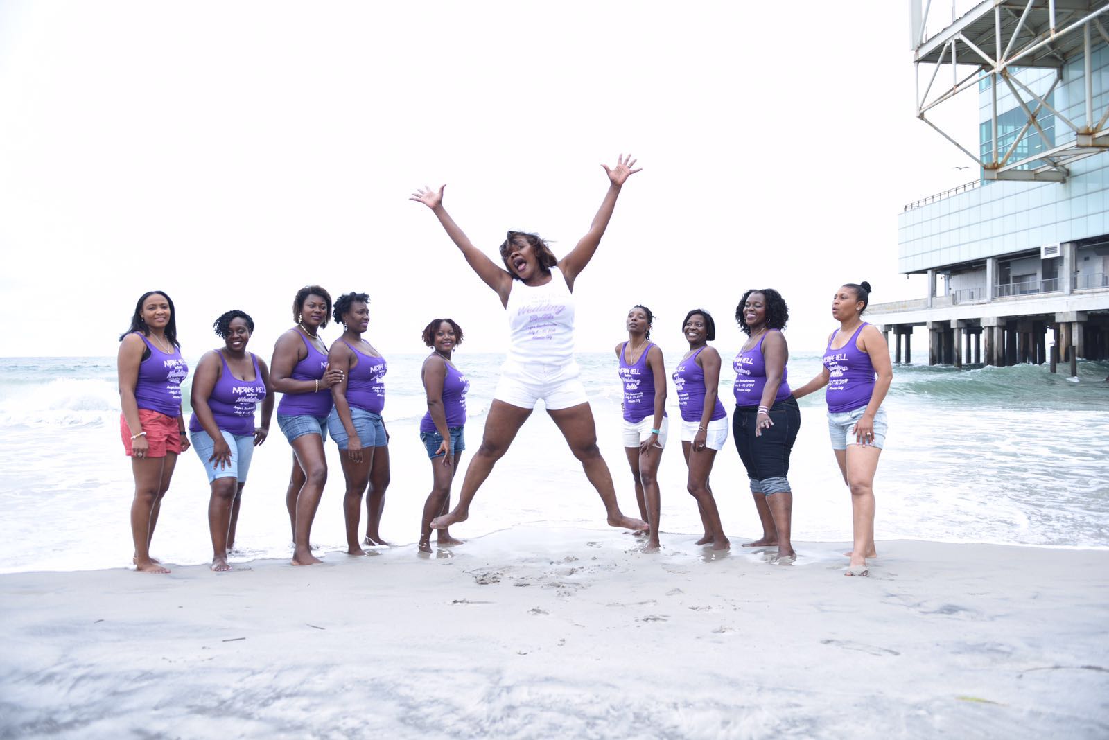 A group at a bachelorette party wearing matching custom t-shirts poses on the beach. The bride is wearing a white shirt, and the others are wearing purple. 