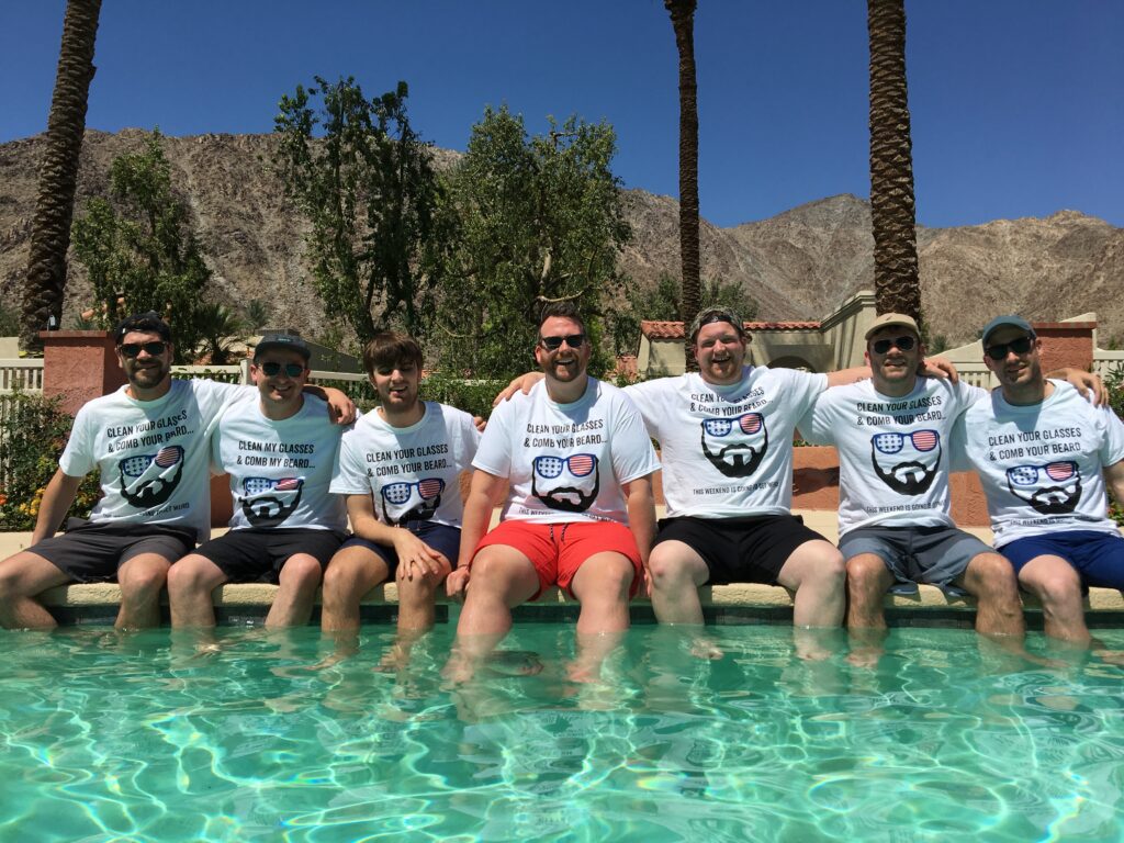 A group of friends at a bachelor party sitting on the edge of a pool and wearing their matching custom tanks. 