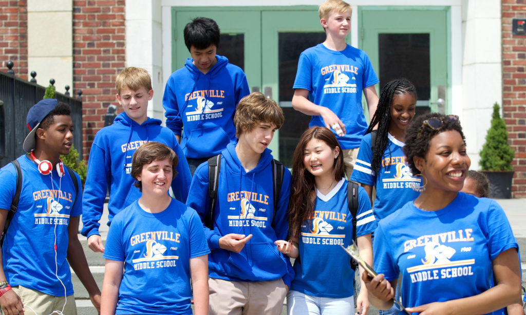 A group of students, accompanied by their teacher, heading out of their school to go on a field trip. They are wearing royal blue t-shirts and hoodies bearing their school name in yellow and white lettering: Greenville Middle School. 