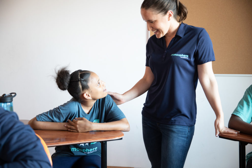 A teacher talks to their student in the classroom while wearing a navy blue polo with "Atmosphere Academy" embroidered on the left chest. 