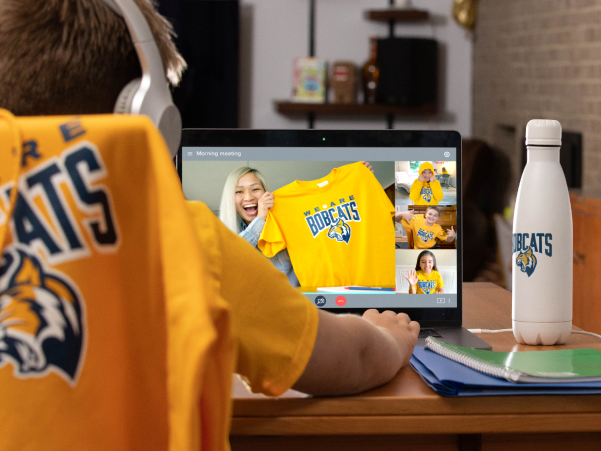 A student sits in their chair wearing headphones in front of their computer. They're wearing a yellow t-shirt with their Bobcats mascot on the front and looking at their computer screen, which shows a video chat with other students showing off their custom school gear. 