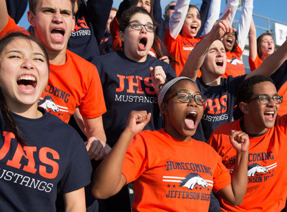 A crowd of students on the bleachers cheer on their team. Their t-shirts are orange and black and show the name of their school and mascot, the Jefferson High School Mustangs. 