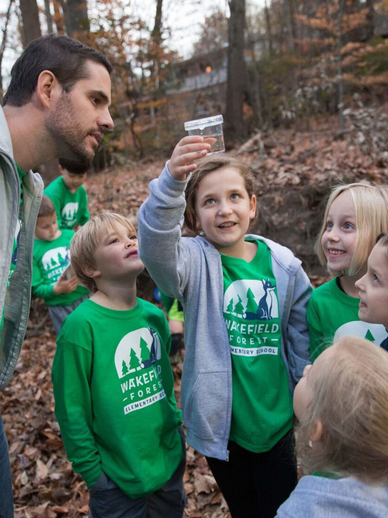 As they explore the woods, a group of students and their teacher wear matching green t-shirts that show their school name, Wakefield Forest Elementary School. They're also wearing matching gray zip hoodies and looking at a specimen inside a plastic cup. 