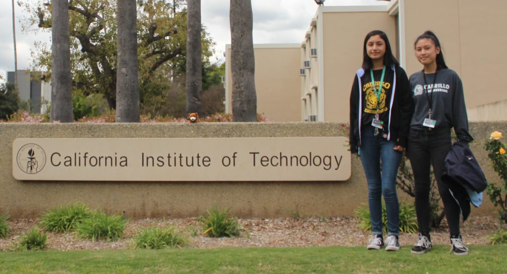 Two young women in custom t-shirts pose in front of the California Institute of Technology sign.