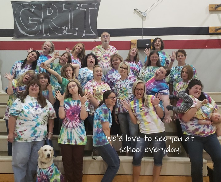 Teachers, staff, one dog, and a kid all stand in the gymnasium showing off their custom tie-dyed shirts.