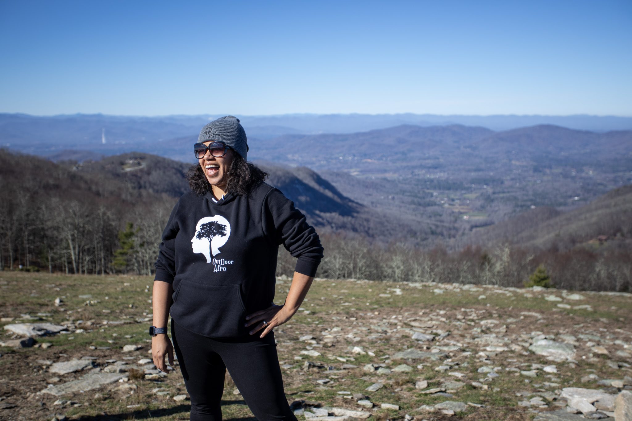 A woman stands on a hilltop smiling wearing her custom Outdoor Afro sweatshirt with their logo that shows the silhouette of a person's head with a tree growing where the brain would be.