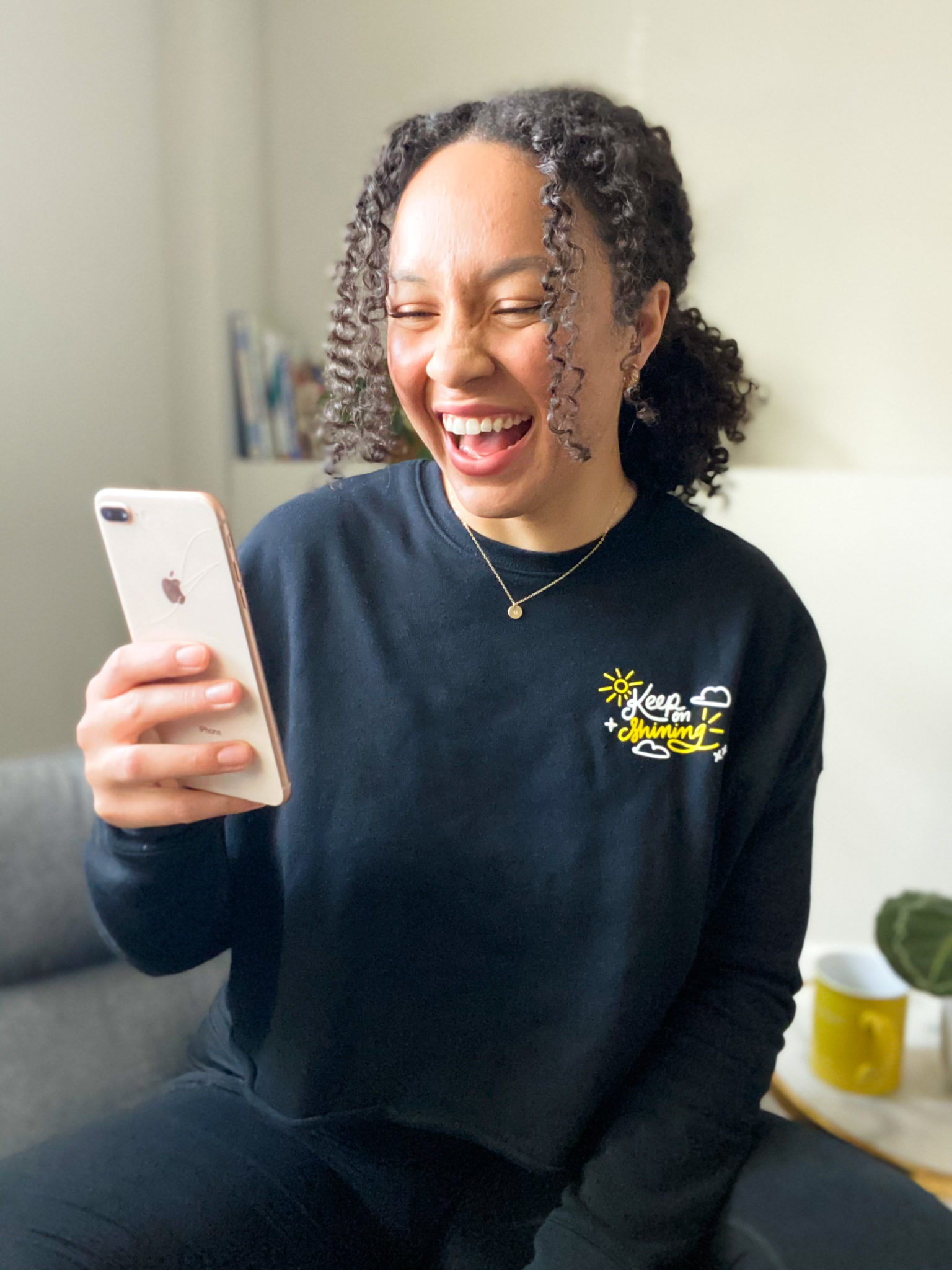 a woman smiles at her phone wearing a black custom sweatshirt with a yellow and white design that says Keep Smiling with a sun and clouds