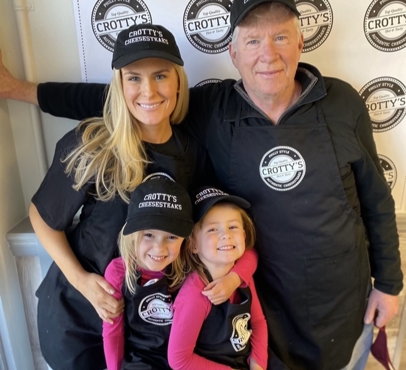 Smiling blonde woman smiling man, and two little girls all wearing Crotty's custom hats, custom t-shirts, and custom aprons stand in front of a wall of Crotty's custom logos.