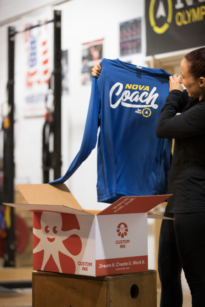 Woman pulling a blue shirt that says COACH up from a Custom Ink box.