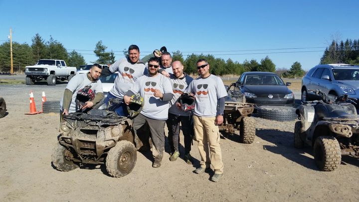 A group of men hang out in matching custom t-shirts with a dirty atv or all terrain vehicle.