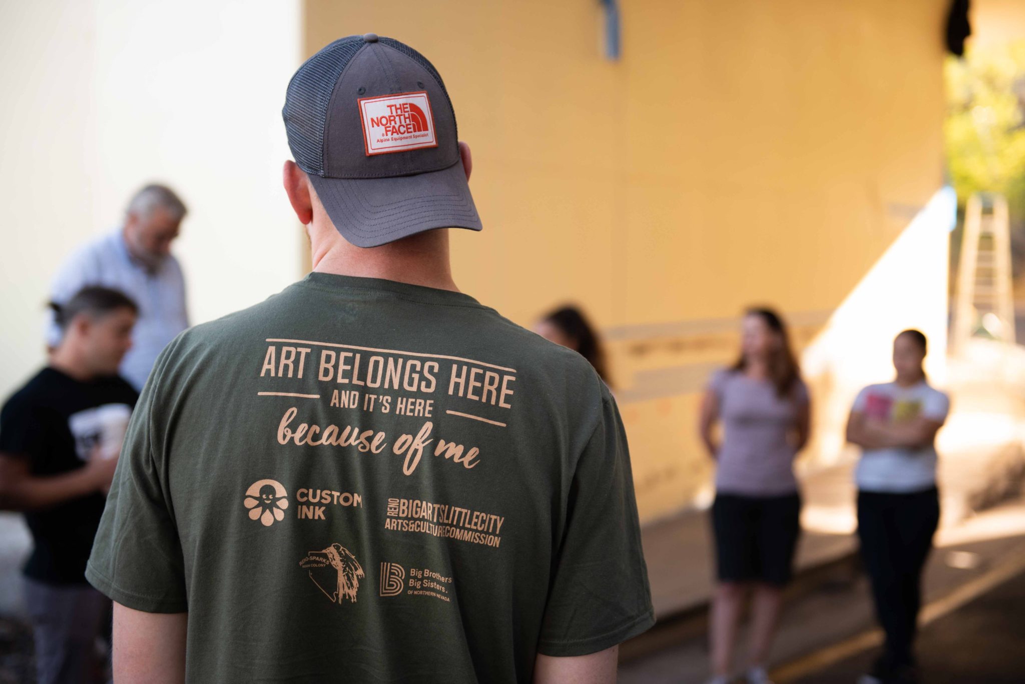 A man from the back wearing a custom t-shirt hat reads "Art Belongs Here and it's here because of me."