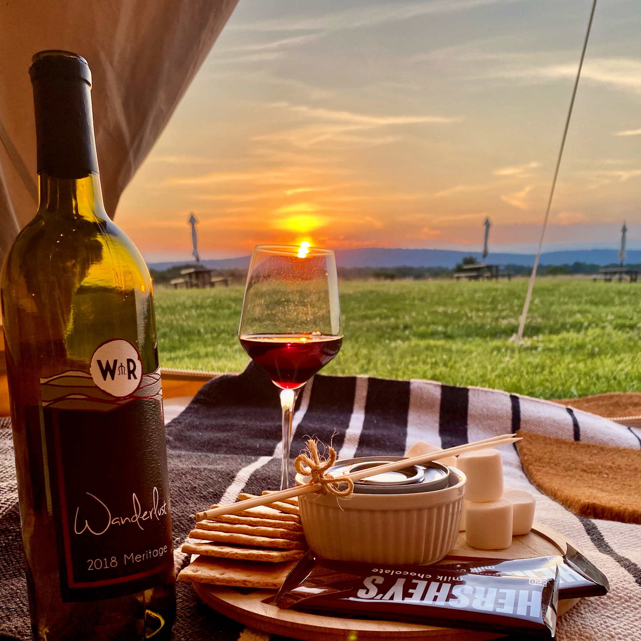 You can see past a wine bottle, glass of wine, and smores kit just inside a tent out to a beautiful sunset over the mountains of Virgnia.