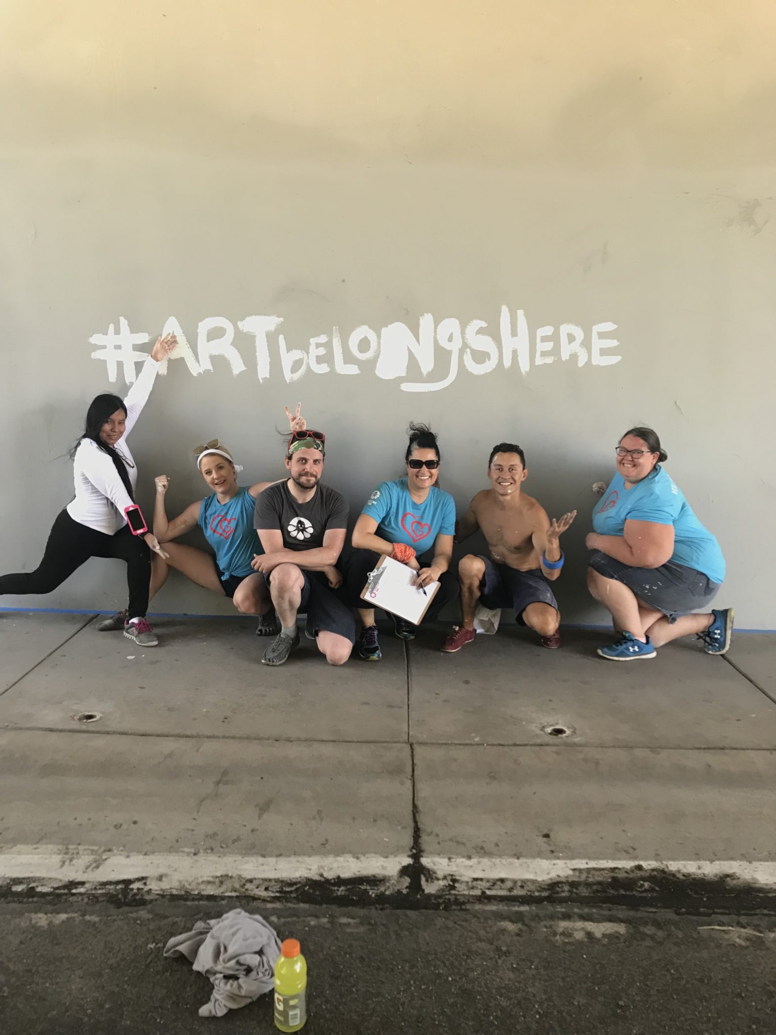 Six people kneel in front of a wall that has the words "Hashtag ART Belongs Here" painted on it.