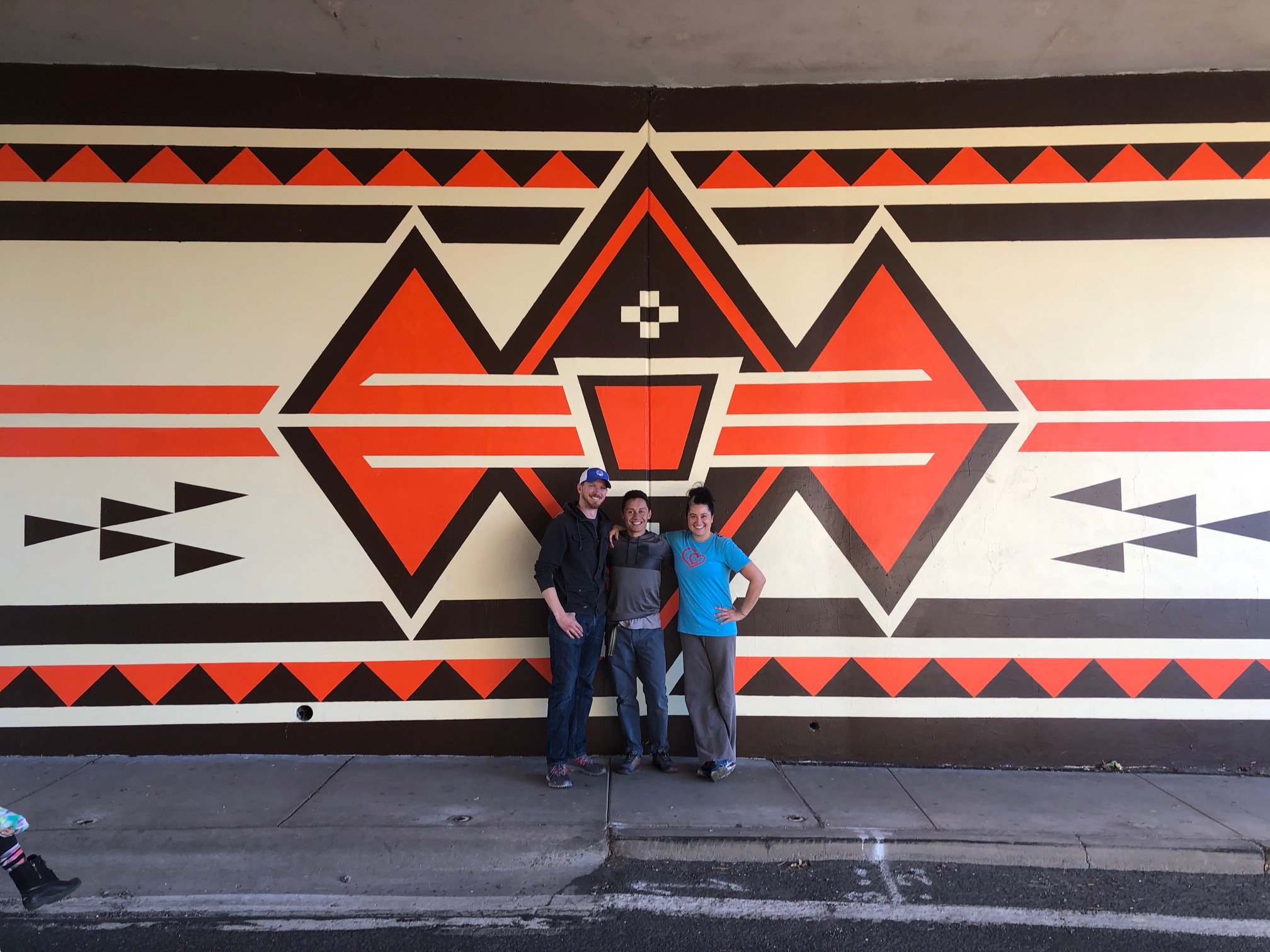 three smiling people pose in front of the completed 2019 mural that is a geometric design with cream, orange, and brown colors.
