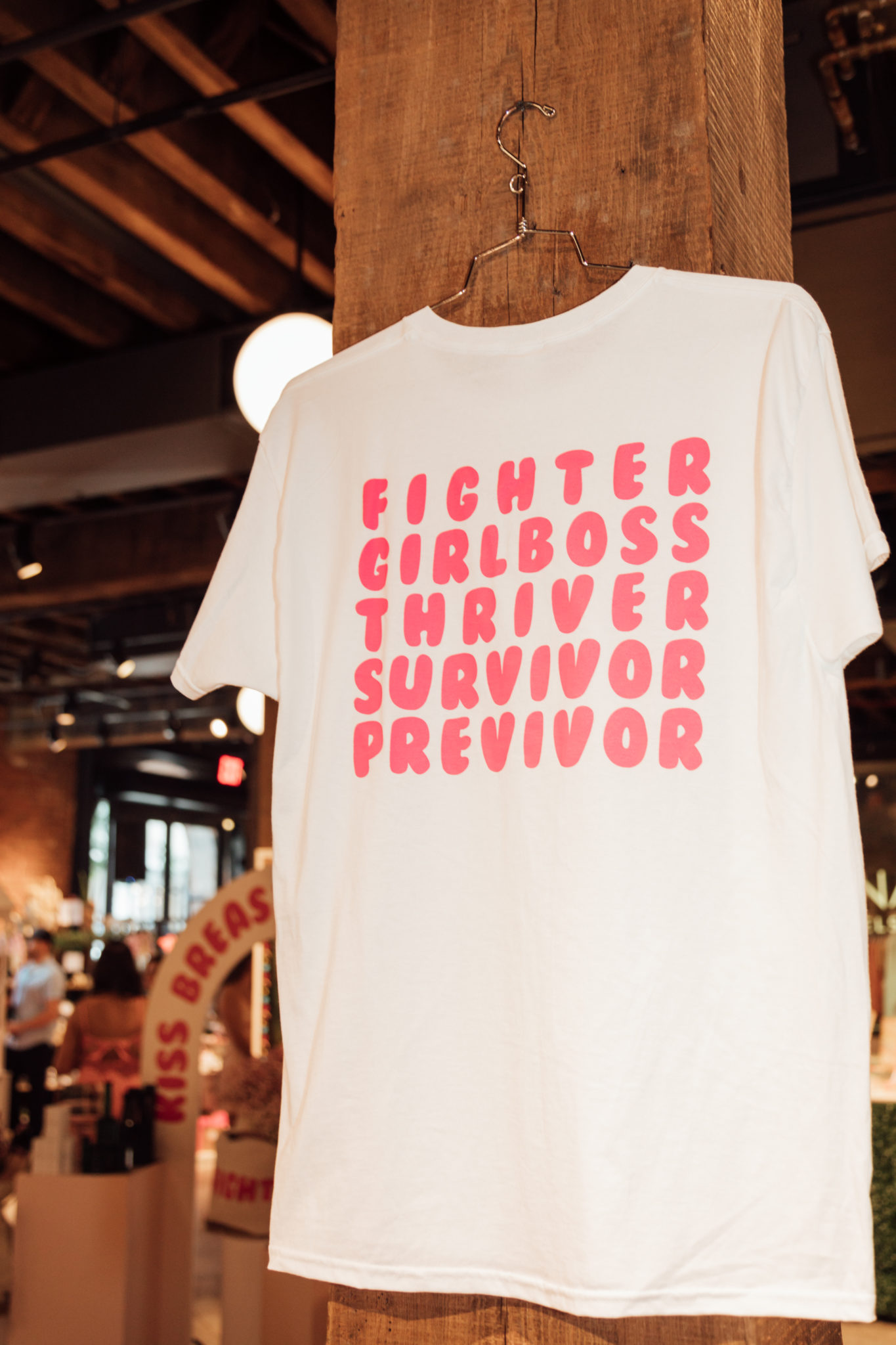A white custom t-shirt, designed for a breast cancer awareness fundraiser. It features the words "fighter" "girl boss" "thriver" "survivor" and "previvor" on it in a bubblegum pink font.