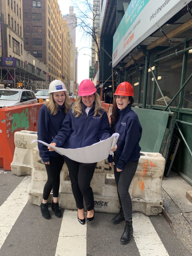 Three women wearing hardhats pose in front of a construction site while they wear matching jackets