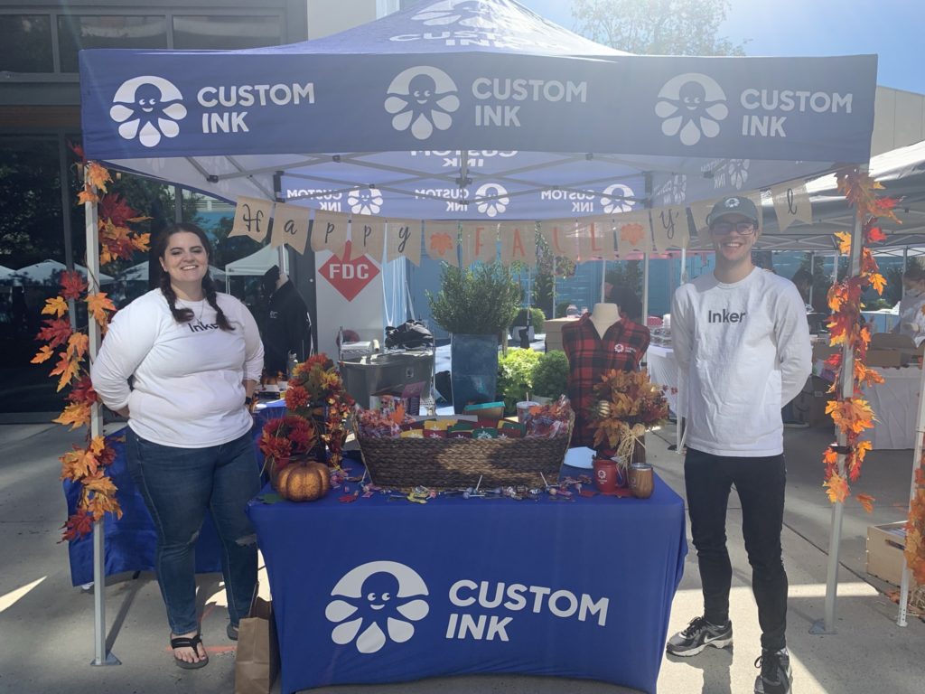 Photo of outdoor Custom Ink Booth setup with two employees standing in front of the table.