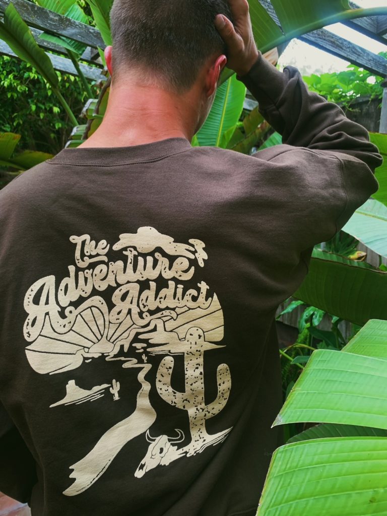Man surrounded by leaves wearing sweatshirt with The Adventure Addict printed on it