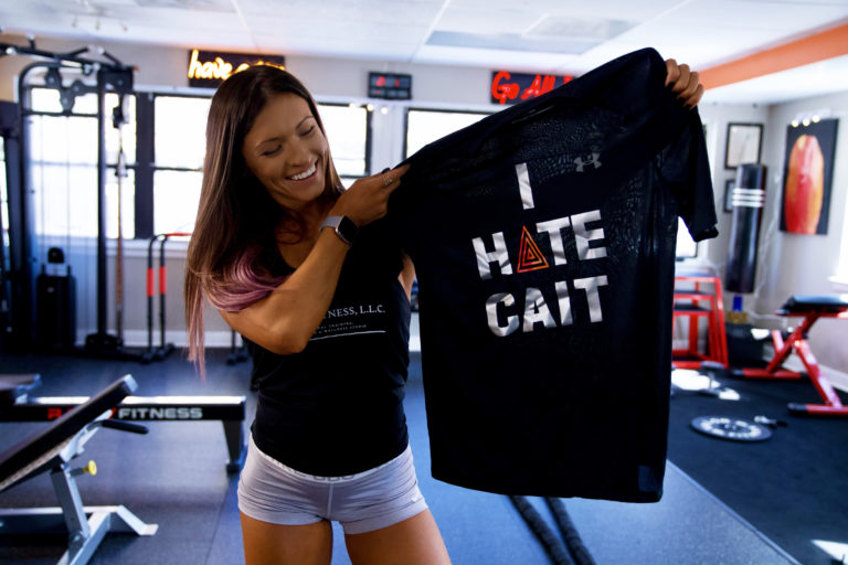 a woman smiles standing in a gym and holding up a black shirt that says "I hate Cait". the A in hate is actually three concentric triangles which is her logo.