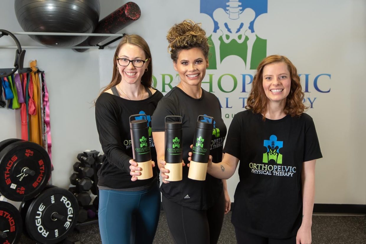 Three women, wearing matching custom shirts that show the Orthopelvic Physical Therapy logo (a puzzle piece with a skeletal pelvis cut out of it) hold water bottles bearing the same logo.