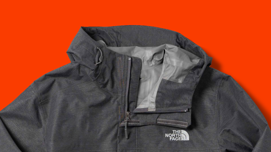 A close-up of a dark grey heather The North Face Waterproof Windbreaker Jacket from the shoulders up. This waterproof fabric is excellent for repelling water and protecting you from the elements. 
