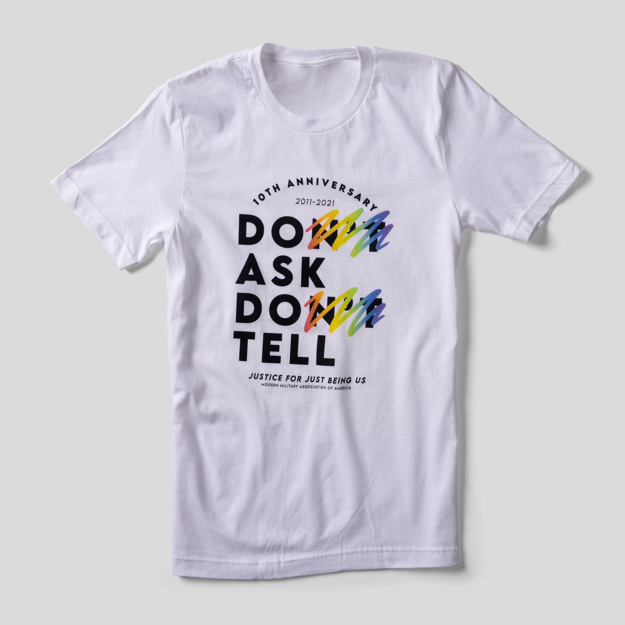 A white t-shirt that reads 10th Anniversary 2011-2021 Don't Ask Don't Tell Justice for Just Being Us. The "n't" parts of "Don't" are crossed out in rainbow scribbles.