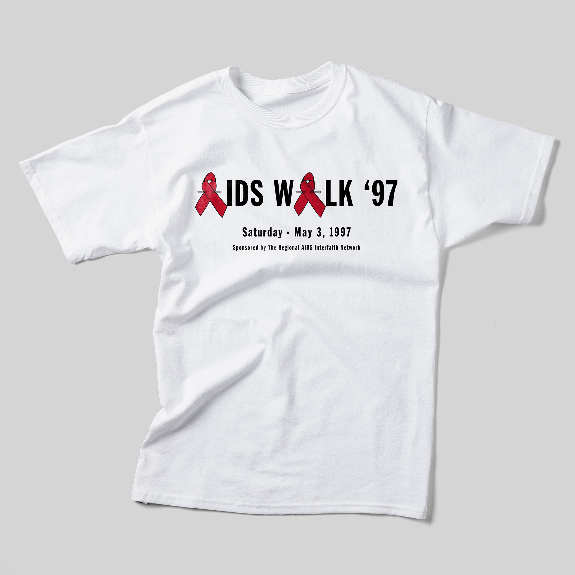 A white t-shirt that reads "AIDS Walk '97. Saturday, May 3, 1997. Sponsored by the Regional AIDS Interfaith Network" in black text. The A's in "AIDS" and "Walk" are red ribbon pins.