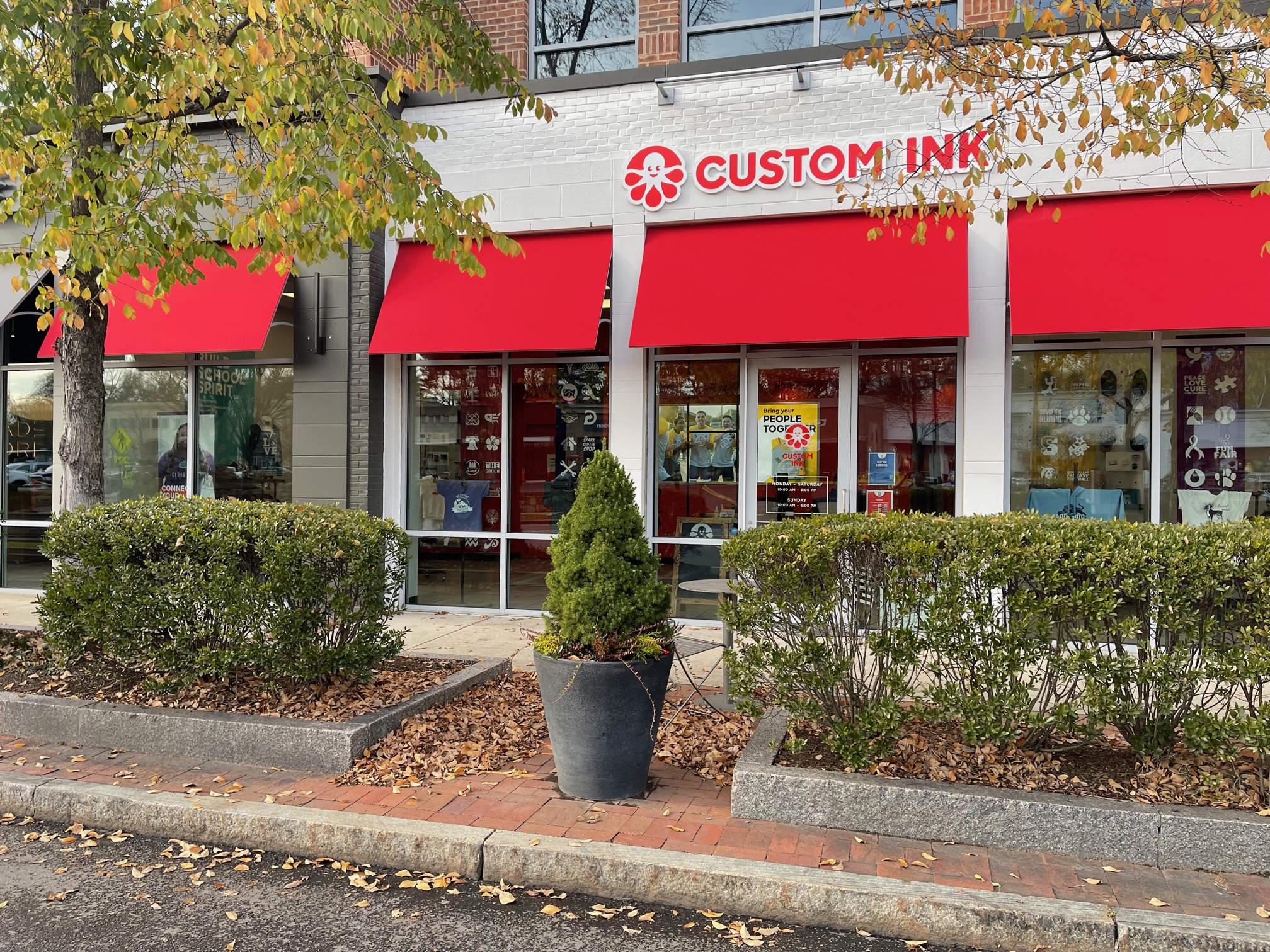 Leafy trees line a street in front of the Custom Ink showroom's facade, featuring the Custom Ink name and logo (a red octopus) and a series of red awnings.
