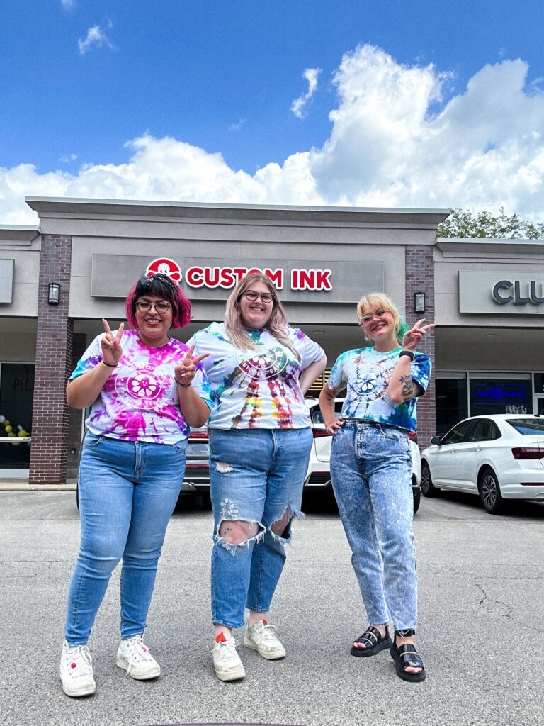 Skylar and two other smiling people wearing tie-dyed shirts in front of the Custom Ink showroom